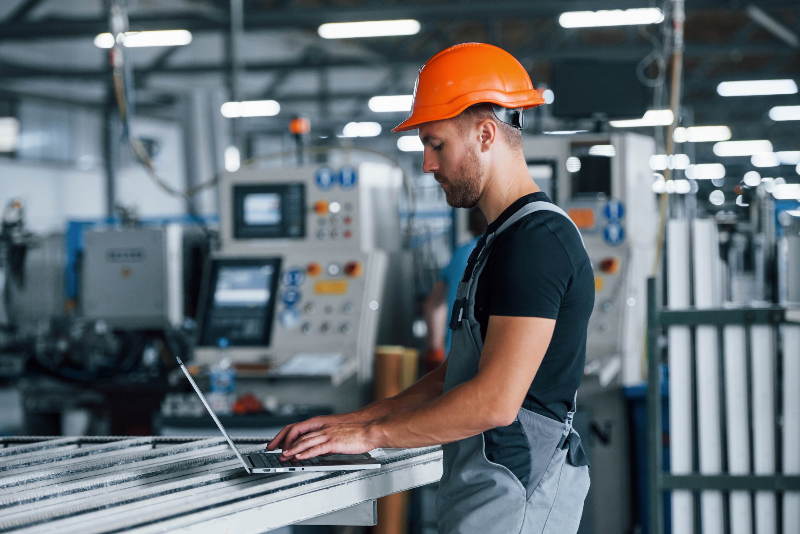 Engineer in an industrial setting wearing an orange hard hat and safety vest, working on a laptop next to machinery