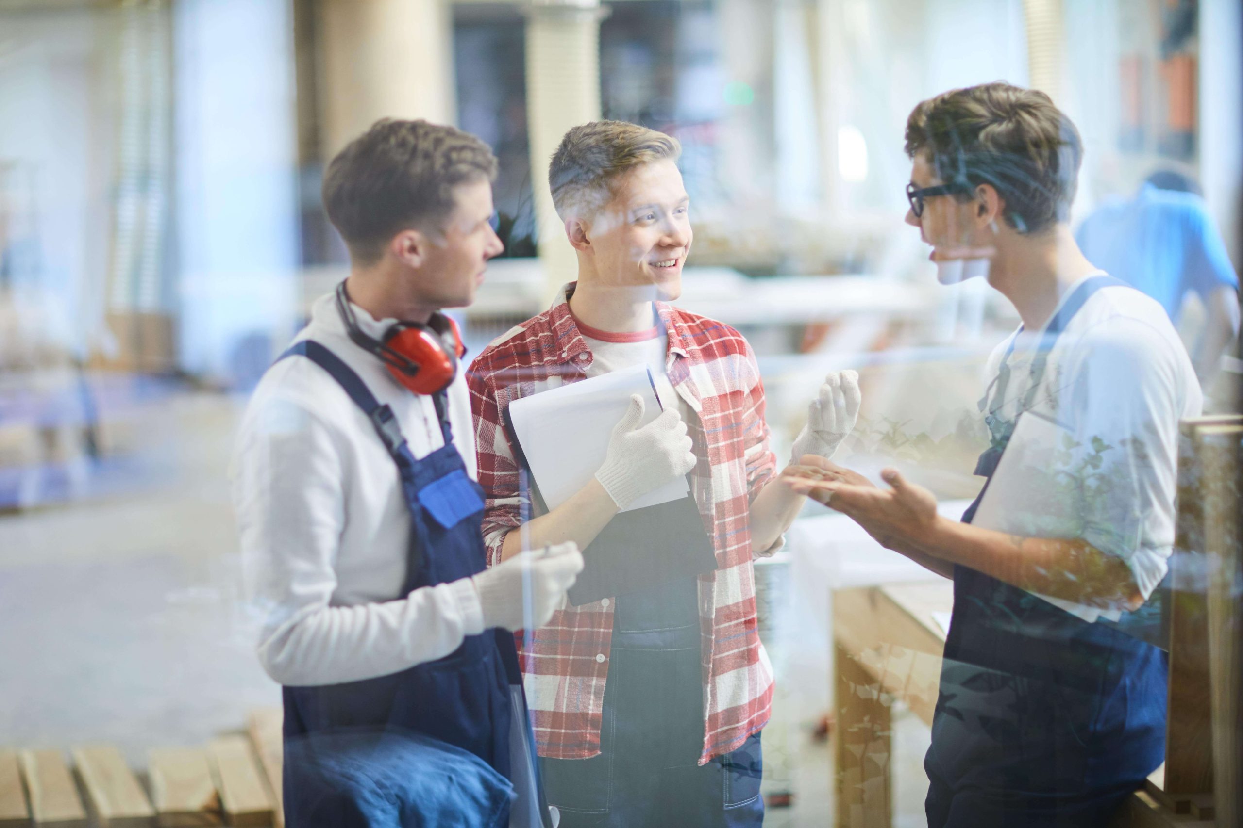 Three workers wearing overalls and gloves having a discussion in a workshop, with one holding a clipboard