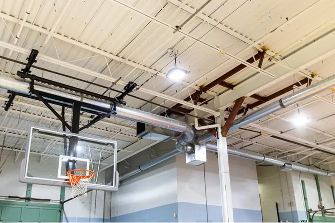 Indoor gymnasium with exposed ceiling showing industrial ductwork and a mounted basketball hoop.