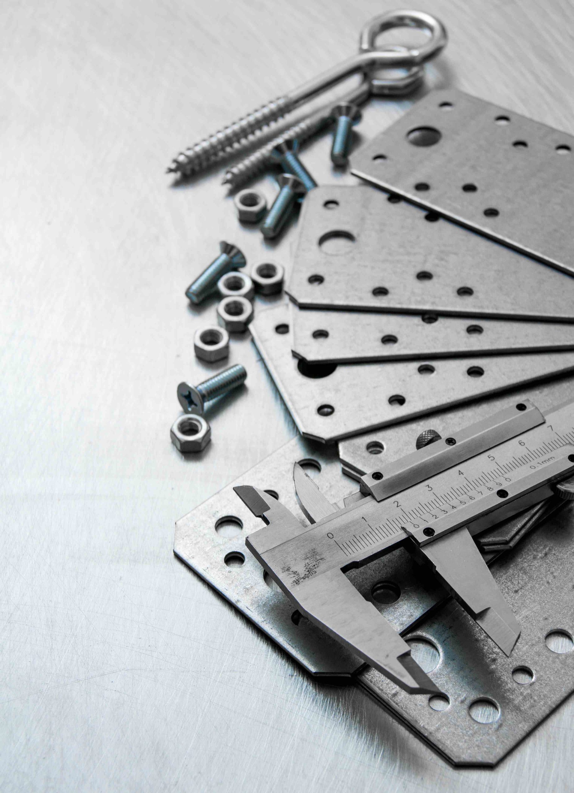 Caliper, blank metal plates, screws, nuts, and bolts arranged on a scratched metal surface.