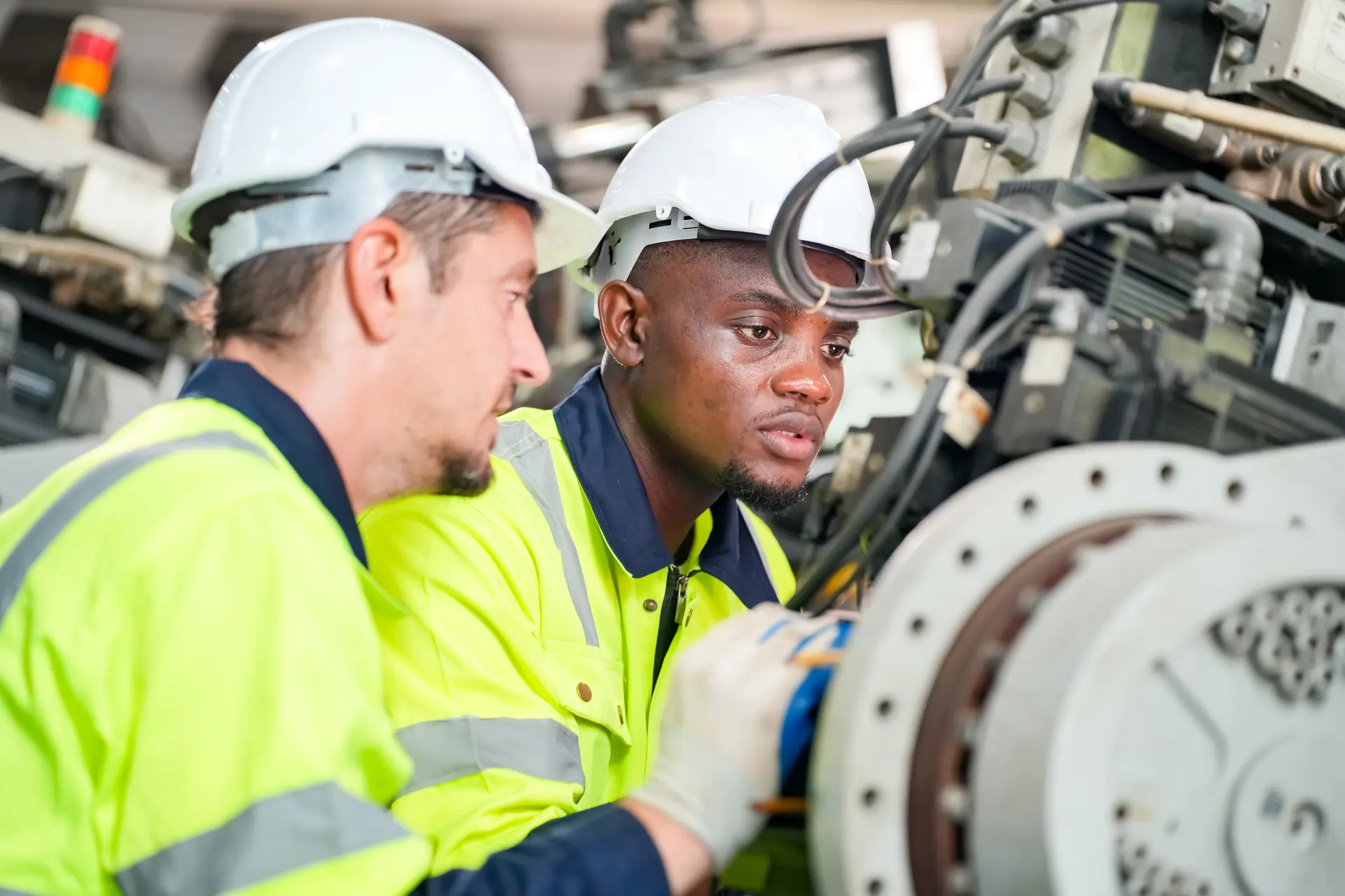 Two industrial workers in high-visibility jackets and hard hats collaborating on machinery maintenance in a manufacturing facility.