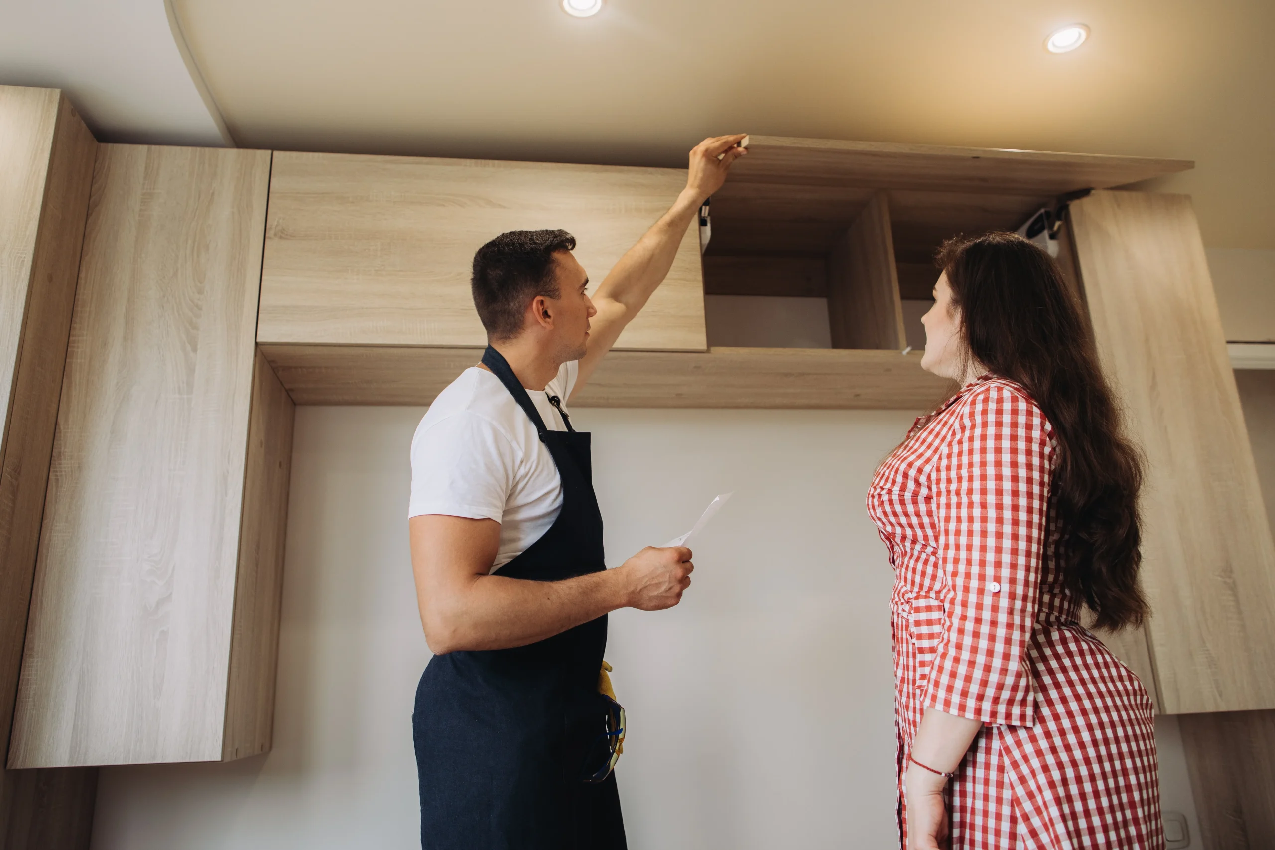 A construction worker in an apron discussing cabinet installation with a client in a modern kitchen space.