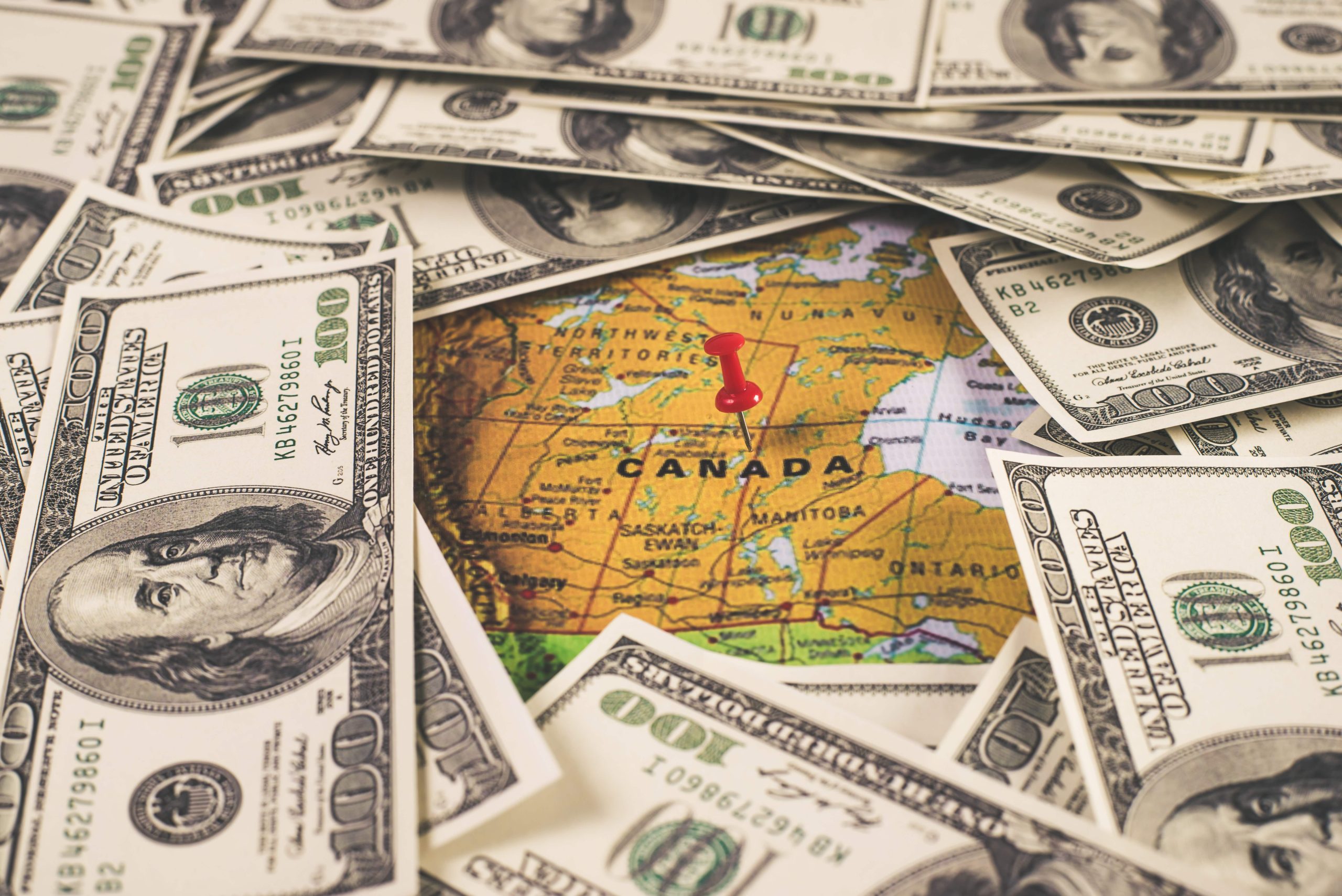 Stack of U.S. dollar bills covering a map of Canada with a red pushpin marking a location, symbolizing potential economic shifts due to trade policies.