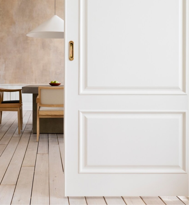 White interior door with elegant gold handle, opening into a spacious room with light-colored wooden flooring and minimalist furniture.