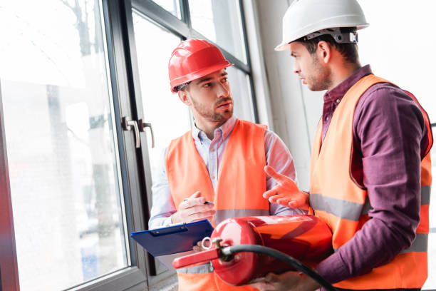 Two fire safety inspectors in hard hats and vests discuss building compliance while reviewing a clipboard and a fire extinguisher near a commercial window.