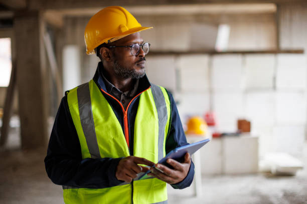 Construction site supervisor in a reflective vest and hard hat reviewing plans on a tablet, emphasizing the role of certified professionals in fire door compliance and building safety.